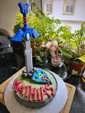 Un gâteau d'anniversaire avec un épée de Zelda plantée dedans (ainsi qu'un bouclier et le nom Mathias en pate à sucre)